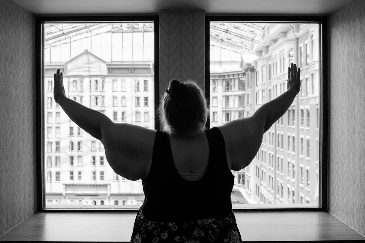 The woman in this photograph by Lindley Ashline has glorious wings on her arms that she lifts towards the ceiling, looking out a window into a large covered open space, in this black-and-white-photograph. She is fat, has long hair pinned into a bun and is wearing a sleeveless dress.
