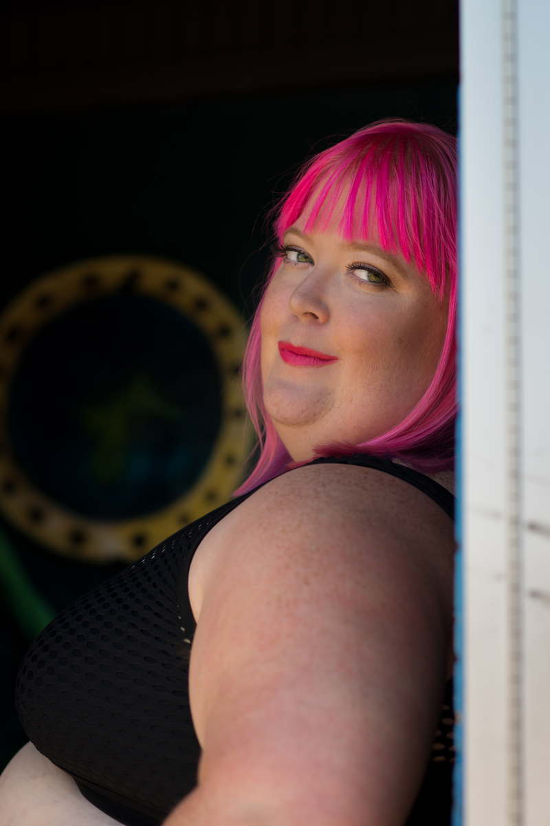 A close-up photo of a fat white person with bright pink hair and lipstick, looking over their shoulder towards the camera. They're wearing a sleeveless black top and leaning against a wall with a nautical-style porthole in the background.